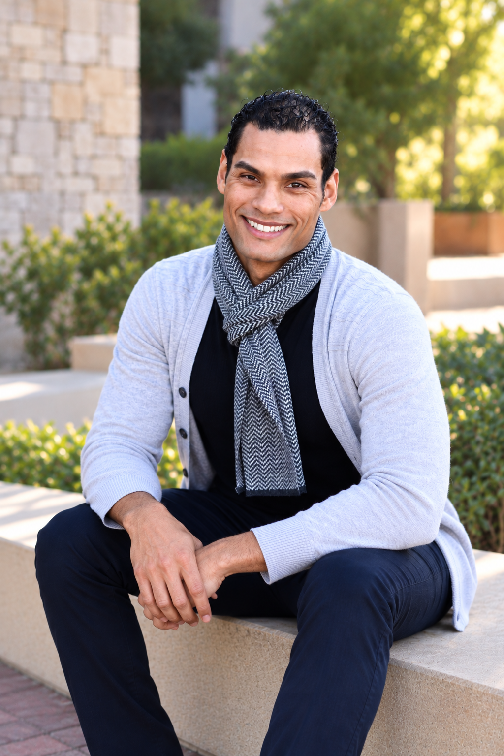 Dr. Samuel Hickson smiling, seated on a stone ledge outdoors in a light gray cardigan, herringbone scarf, and navy trousers
